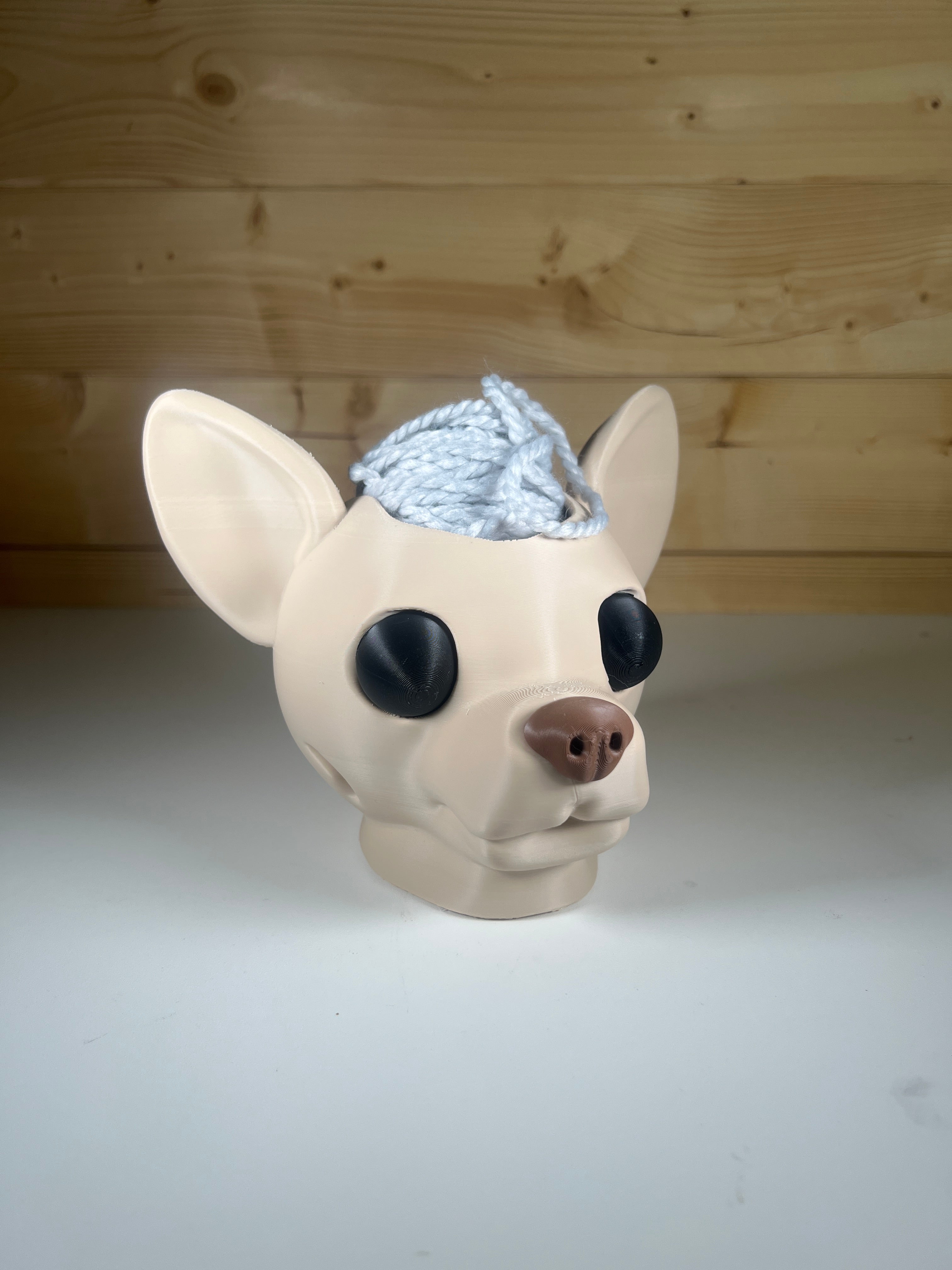 Tan Chihuahua dog head yarn ball holder with white yarn sitting on a white desk with a wooden background.